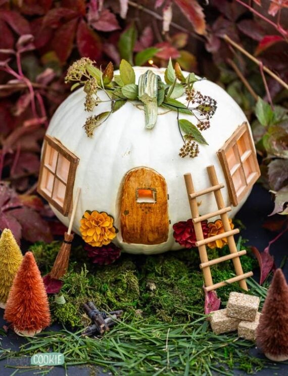 White pumpkin carved into a whimsical fairy house with windows, door and greenery.