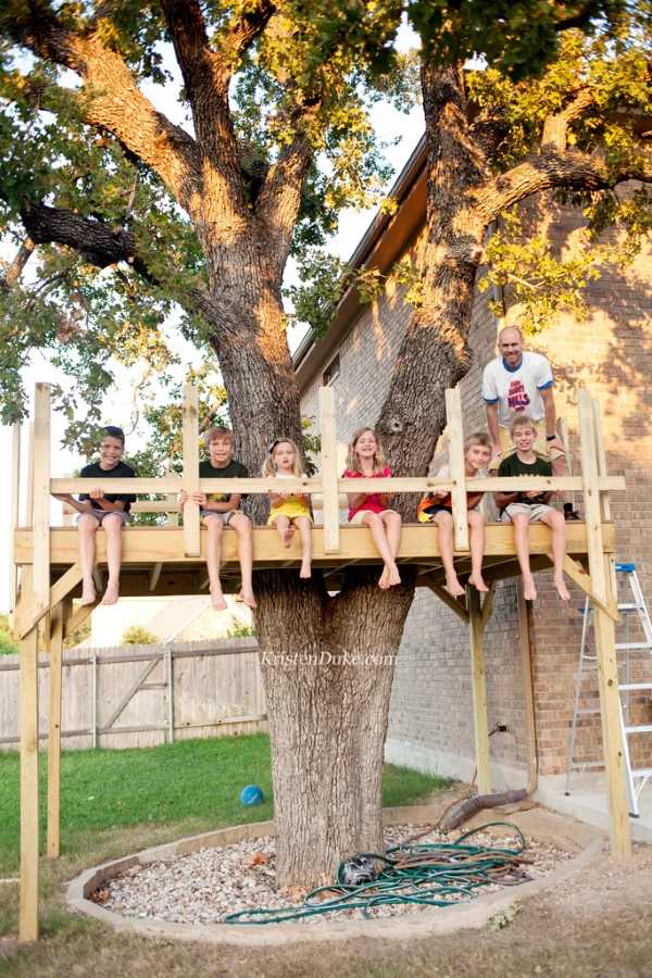 Kids sit on a large DIY tree house platform built around a tree in the backyard.