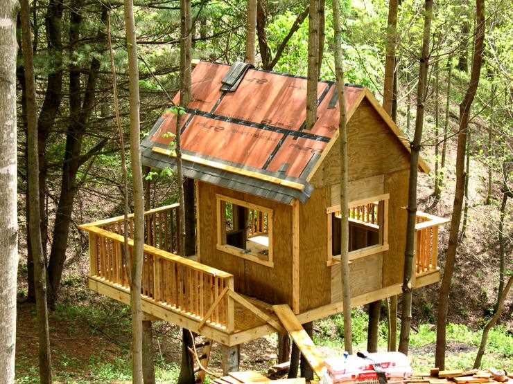 DIY wooden treehouse with red metal roof and wraparound deck built among tall trees.