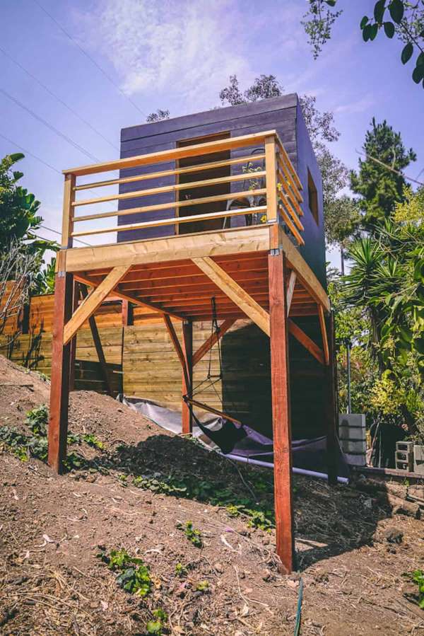 Modern elevated DIY tree house built on wooden poles with balcony deck and hammock below.